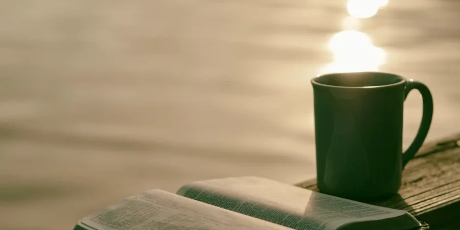 Une bible ouverte devant un plan d'eau au soleil couchnat avec une tasse en céramique verte à côté